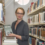 Professor Claudia Weber mit Ihrem Buch "Der Pakt" vor Bibliotheksregalen